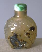 A snuff bottle with raised relief carvings of a man, trees and a monkey (?). On top is a jadeite stopper.