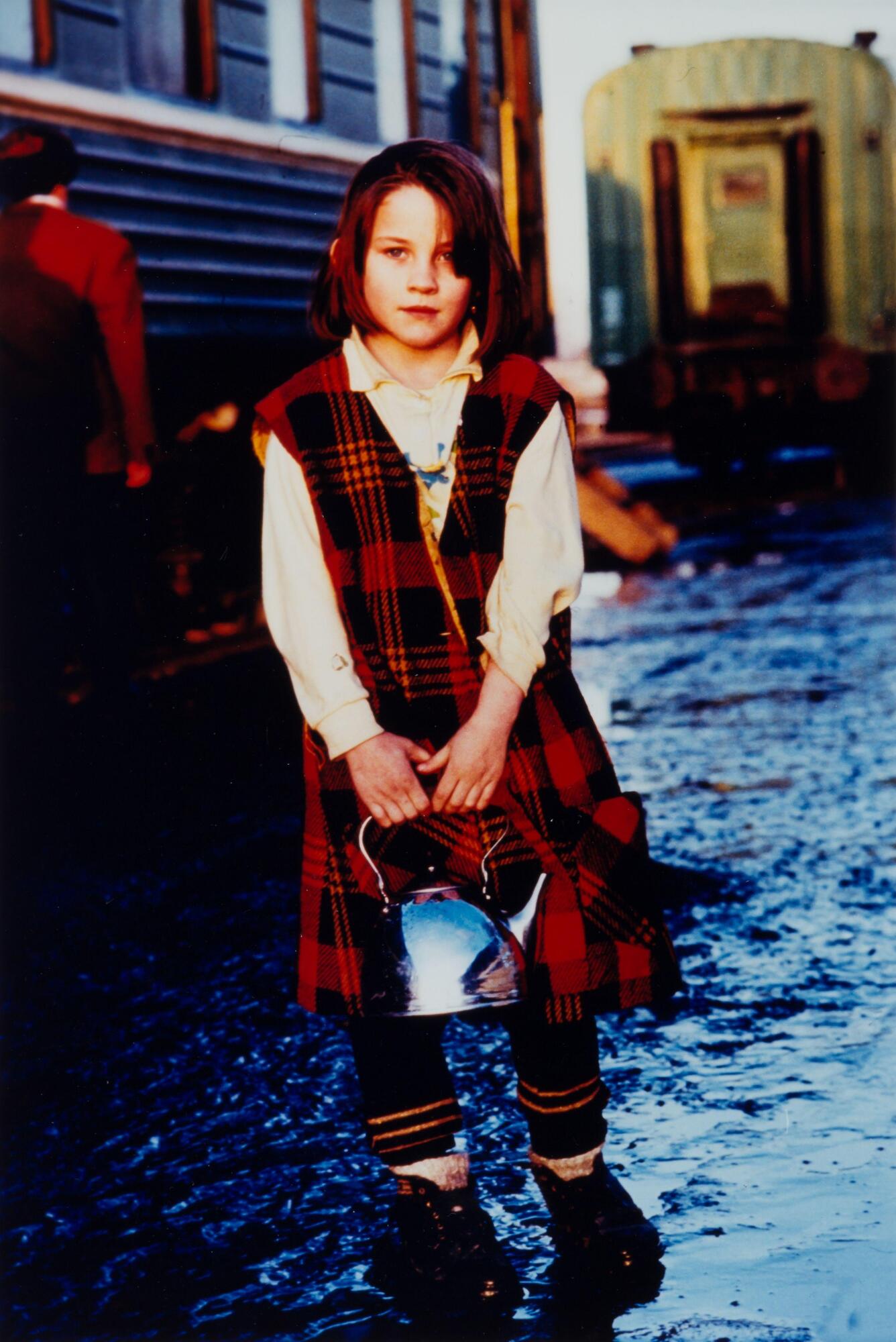A girl on the street in a red plaid uniform holding a silver teapot.