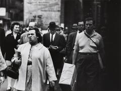 Image of a large group of people walking in an urban setting, all in the same direction. The woman in the foreground holds a handbag and a newspaper. A man on her left carries a large parcel wrapped in paper and string.