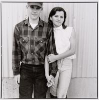 A boy and a girl standing against a metal wall holding hands. She is wearing a horse riding outfit and he is wearing a baseball cap and flannel shirt.