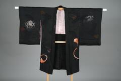 <p>long black haori with interwoven fan and scroll motifs with embroidered bells, ribbons, hand drums, and braided tie-string motifs and three family crests on the backside with a light pink inner lining with interwoven floral chrysanthemum and peony motifs.</p>
