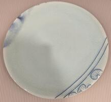 A plate from the center of the piece. The plate is mostly white with two thin blue lines and scrolling designs in the lower right and the faint outline of a man&#39;s mouth and chin in the upper left.&nbsp;