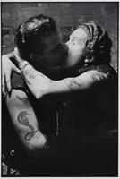 A photograph of two men kissing. The man on the right wraps his arms around the other. Each have tattoos on their arms and wear cut-off vests.