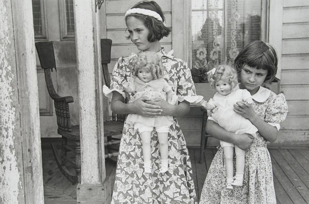 Two girls in floral dresses stand on a wooden front porch holding matching dolls.  Both girls stare inquisitively to their right.  Behind them sits a rocking chair and windowsill with lace curtains and a flower box.