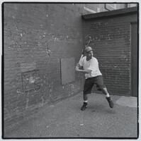A photograph of a young man playing Wiffleball. He stands near the corner of an exterior brick enclosure, leaning back on one foot, preparing to hit the ball.