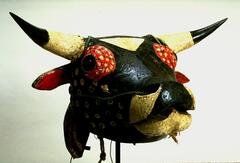 Naturalistic cow’s head of carved from wood with eyes cut from the bottoms of glass bottles. Natural cow horns are attached. Head is predominantly black with white triangle on forehead, red pigment surrounding eyes, red in interior of ears, white chin, and white dots on cheeks and in red ground around eyes. Horns are white, tipped in black; a fiber cord runs through its nostrils and around back of head.<br />