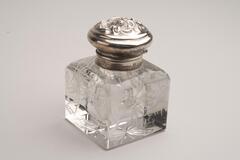 A square, crystal inkwell with rounded edges.  The base is engraved with flowers and ivy. The sterling silver lid also has a flower on the top.