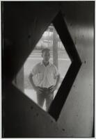 Photograph of a man taken through a diamond-shaped door window. He stands outside, arms akimbo.