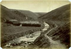 This photograph depicts a river stretching through a valley. The river water cascades over rocks in the foreground. A small road parallels the right back of the river. A small bridge with a figure connects the two sides of the valley in the middleground.