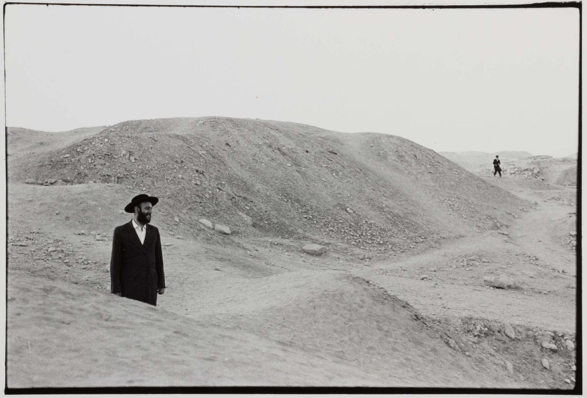 A man in hassidic clothing standing in the middle of a sand pile. There is another man farther back in the distance.