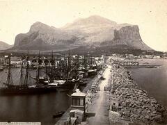 This photograph depicts a view of the harbor of Palermo, a stone pier running through the frame and a fleet of wooden sail ships at their moorings. In the background rises the mountainous land formation of Monte Pellegrino.