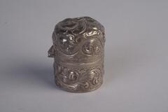A cylindrical sterling silver inkwell. The outer casing is decorated with a scroll design and opens halfway down the inkwell.