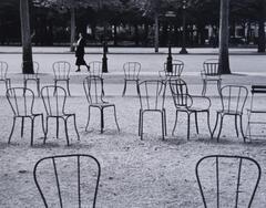 Photograph of metal chairs in a park adjacent to the Champs-Élysées Avenue in Paris. In the background, a woman walks by.