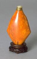 A baltic amber snuff bottle that is shaped like a diamond. On the top of the snuff bottle is a mother of pearl stopper in a brass collar.