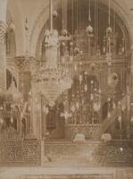 This photograph shows an interior view of a highly decorated religious building. A chandelier and various other hanging lamps fill the upper register of the image. Other architectural elements in the picture, including the floor, the walls, a railing, the altar, and other shrines, all appear covered in intricate foliate designs.   