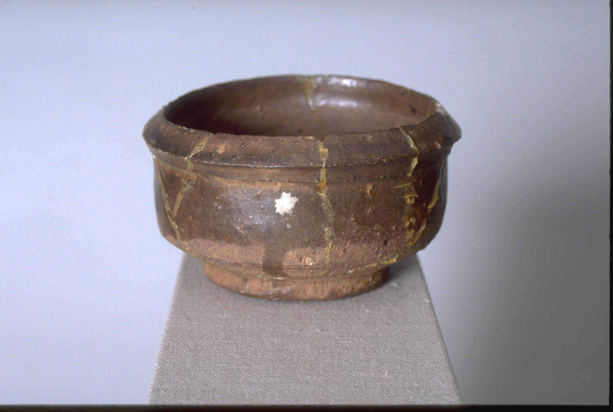 A small, thickly potted, squat, brown stoneware bowl on a wide footring and everted rim.  It has concave articulation, and brown glaze covering half the exterior and the entire interior body, with repairs.