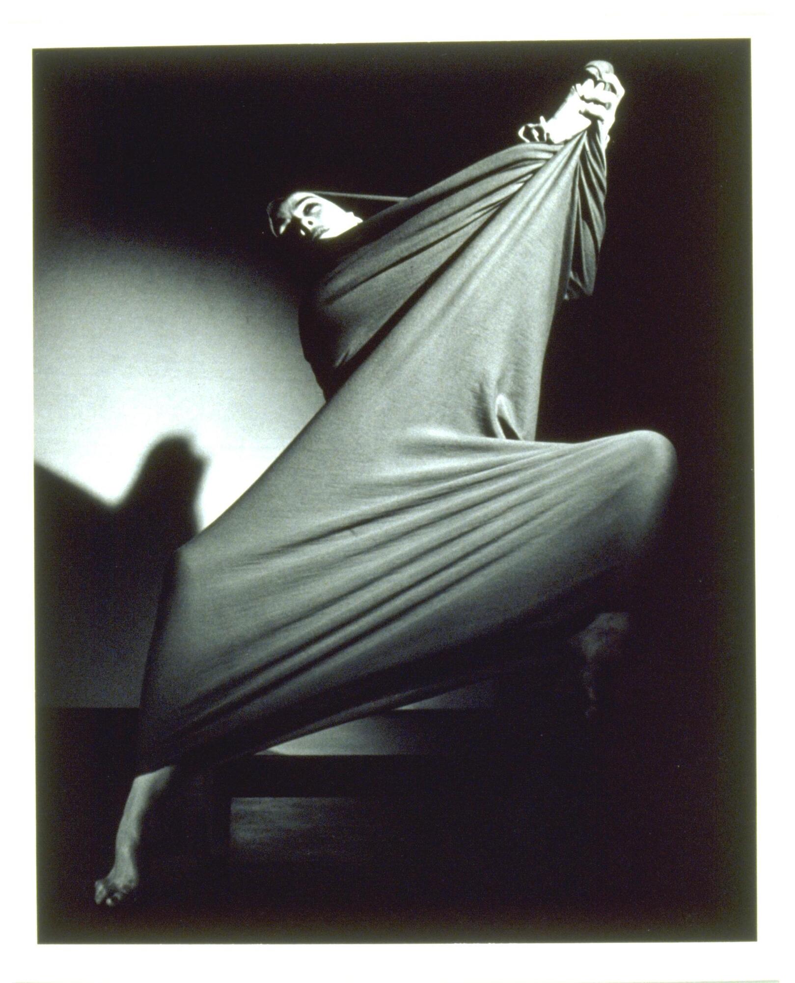 A photograph of a woman dancing. She lifts her arms up toward the right of the frame, her legs creating a diagonal across the bottom of the frame. 