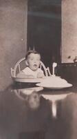 A small child in a high chair, seated behind what appears to be a cake with a single candle in it. The child&#39;s mouth is open as they gaze up towards the upper left of the image.