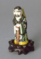 A snuff bottle in the form of a bearded male figure. He is wearing a black robe with white flowers and a yellow and green trim. He is holding a brown staff with both hands.