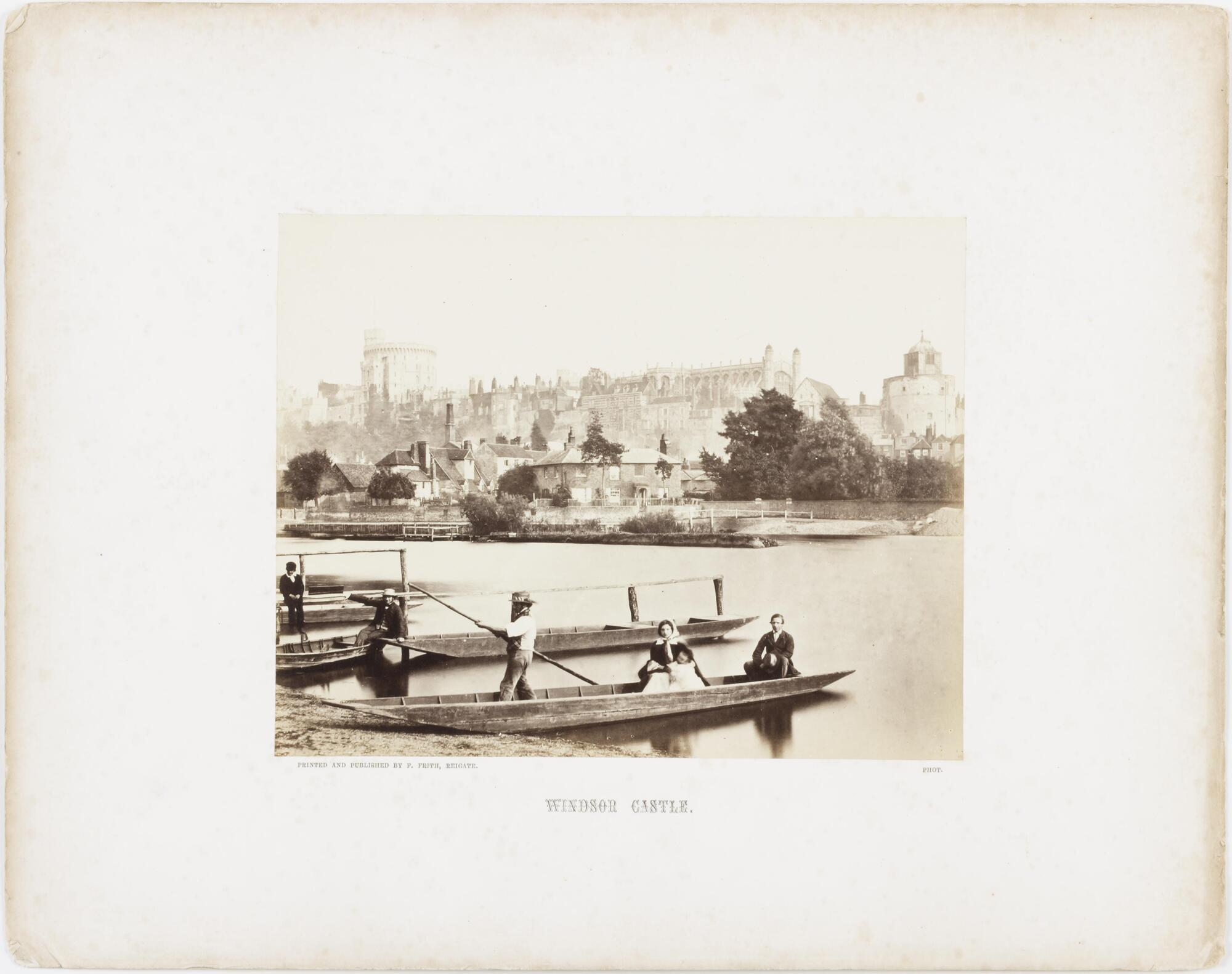 View of several row boats with passengers moored on the edge of a body water across from a town. 