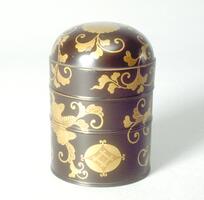 A small cylinder with rounded top and several grooves going up the height of the object. Golden lacquered floral patterns cover the surface with one crysanthemum crest on the top center of the lid and a geometric crest near the bottom. This is the larger of two containers. Part of a bridal trousseau.