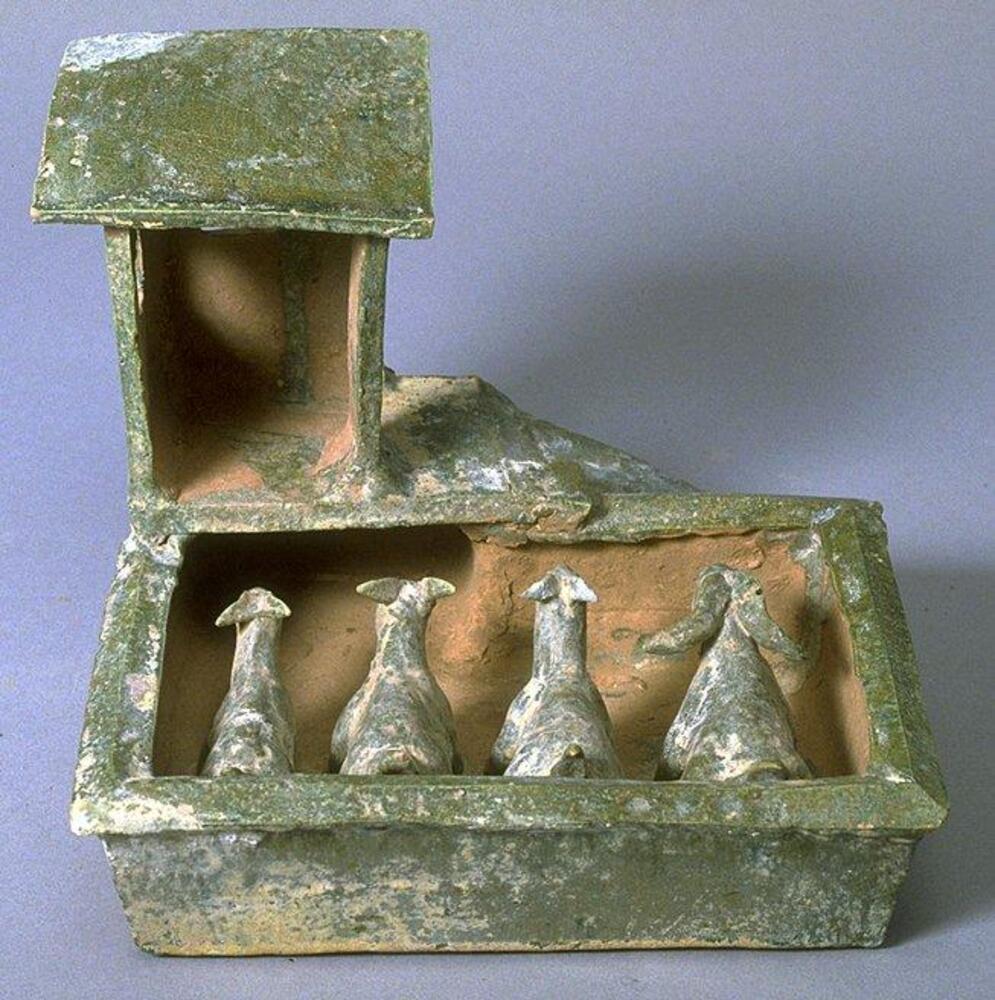 A red earthenware ceramic model of a goat pen, containing one ram and three ewes, with a small shed over the pen with stairs.  The exterior and goats are covered in a green lead glaze with iridescence and calcification.