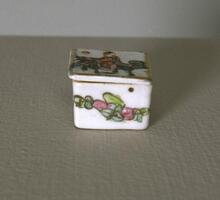 A white square rouge box with a painting of a floral design and a lid with a painting of a man on a horse.