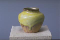 This small vase has a fairly wide mouth and a gently tapering profile. The upper 2/3 of the vessel has a thick creamy yellow glaze with traces of blue. The glaze has drip marks that run down the face of the vase.
