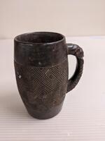 Carved cup with a curved handle decorated with a human face. The body of the cup has decorated with geometric designs.&nbsp;