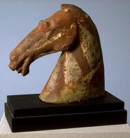A gray earthenware head from a horse sculpture, its strong muscular neck holding its narrow head high. It is vividly sculpted to show the musculature of the horse's face with flaring nostrils and open mouth showing its tongue. It has deep set and bulging eyes, ears pointing forward and a flowing mane. The head is covered with red pigment and with painted white pigment horse trappings. 