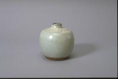 This is a small, globular stoneware jar with a slightly tapered base and a narrow, tapering, short neck. A raised, wavy line decoration encircles the shoulder. The jar is covered in a pale, mottled, blue-green glaze. 