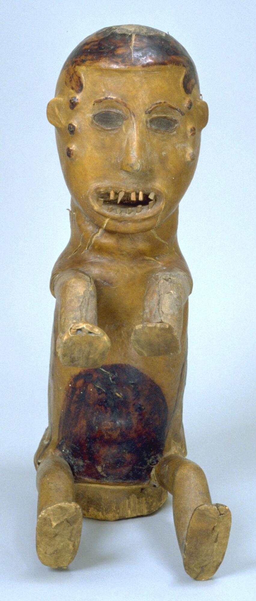 A wooden crest mask covered with antelope skin in the form of a human figure with a columnar body and four outstretched limbs. The center of the figure's abdomen is dyed a dark brown, while the rest of the skin is light in color. On each side of the face are three dark circles in a vertical line. 