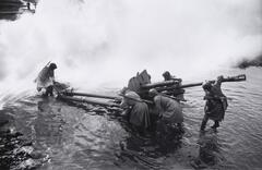 A group of soldiers drag a piece of artillery through shallow water. 