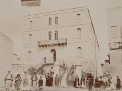A large, four-story stone building with numerous male figures standing either on the front balcony or in single file just out front. On the right-hand side of the photograph, two signs on a nearby building, written in English, read “Hotel” and “Hotel of Nazareth.”