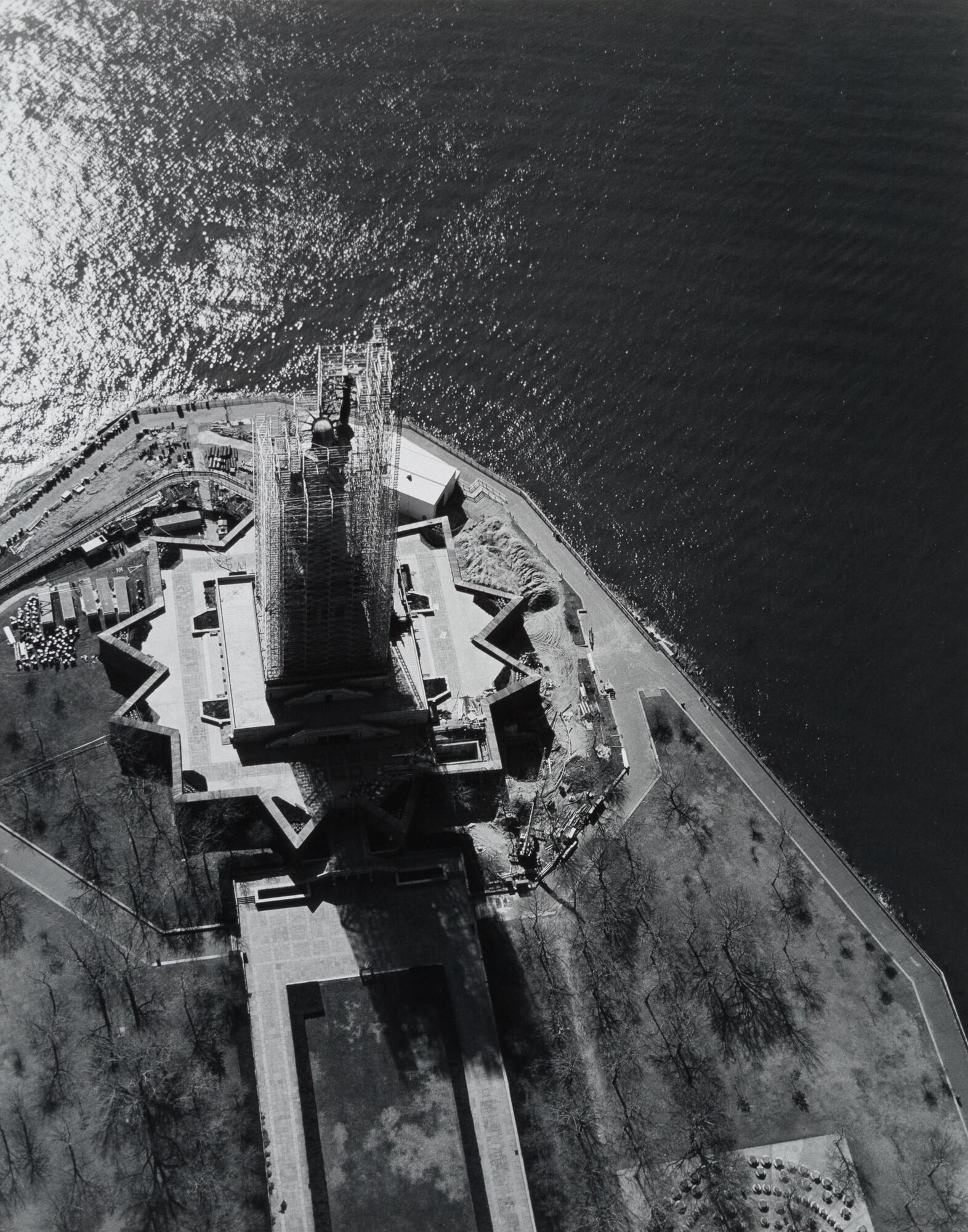 This photograph depicts an aerial view of a statue surrounded by construction scaffolding on an island surrounded by water. 