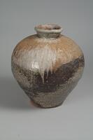 This large jar is wider in the top portion, narrowing towards the base. It has a short neck that flares outwards. The natural glaze creates a range of browns and grays.