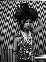 Black and white photograph of a woman with two large bundles resting on her head, which she holds in place with her left hand. She stands in front of vibrant prints with geometric designs and her clothing consists of numerous patterns and textures.&nbsp;