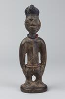 Standing human figure on a round base with hands at the sides. The abdomen is marked by triangular grooves and around the waist is a string of beads. There is also a string of beads around the neck, while the forehead has four vertical, incised grooves and the chin has three grooves. This figure also has closed eyes, not seen often on Yoruba twin figures. The hair is in the shape of a knob with vertical incised grooves. 