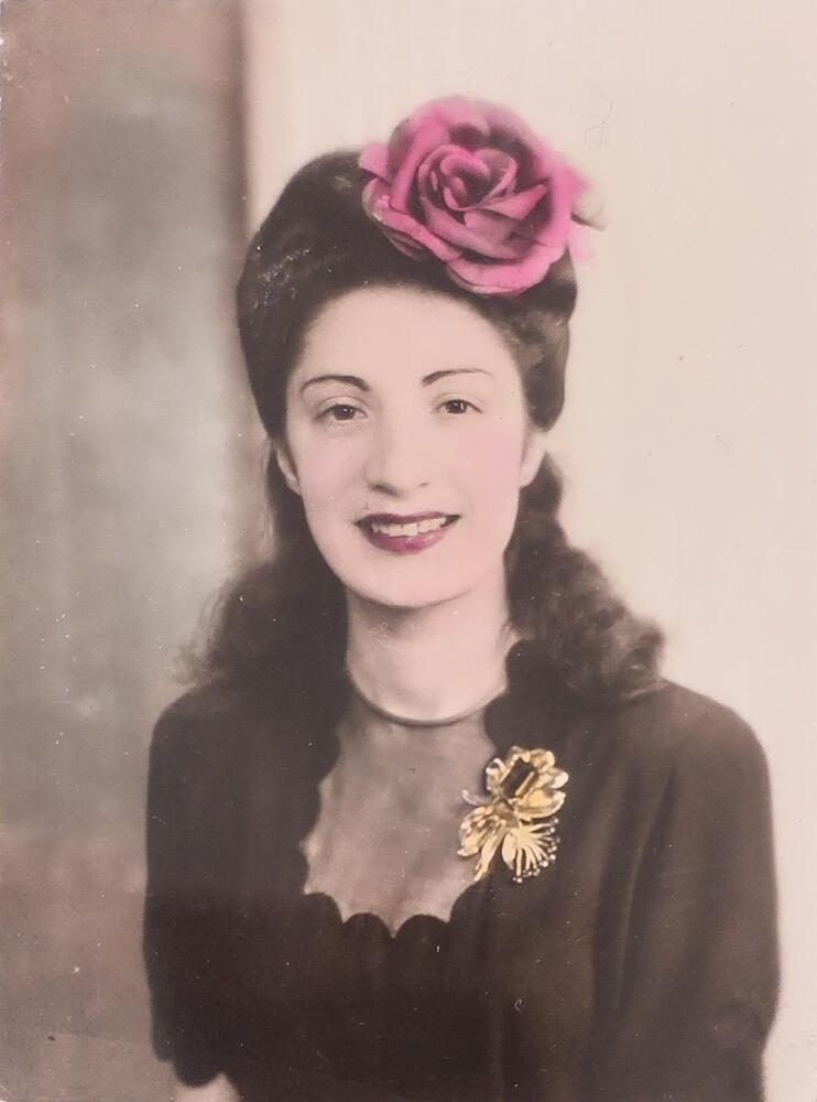 What appears to be a colorized portrait of a woman with a large pink flower in her hair. She is directly facing the camera and smiling with a large gold brooch near her left shoulder.