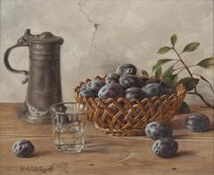 A still life on a wood table with a white cracked wall behind. On the table are plums, some in a wicker basket and a few on the table. There is a water glass half full and a metal tankard. Behind the basket is a branch with green oval leaves.