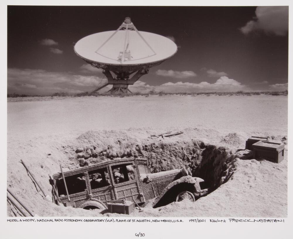 This photograph depicts a view of an archaeological excavation of a car buried in a desert. In the background is the large radio dish of an observatory. 