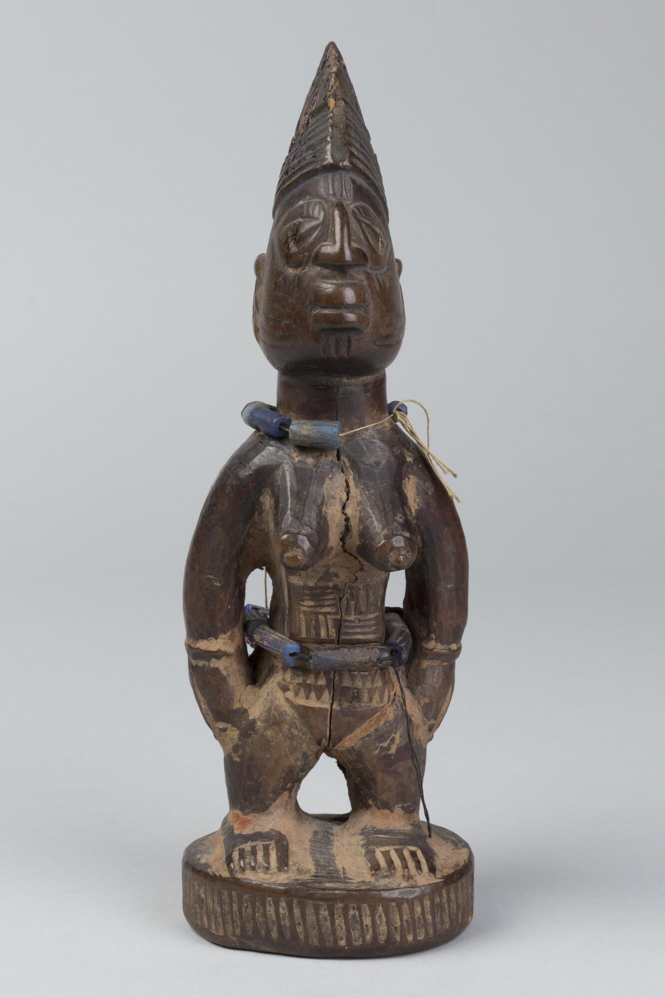 Standing female figure on a round base with hands at sides and prominent breasts. There are geometric patterns with lines and triangles on the abdomen as well as beads strung around the waist. Around the neck is another string of beads and the cheeks have incised marks, four on one cheek and two on the other. The hair is in a conical shape with horizontal grooves along the lower half. 