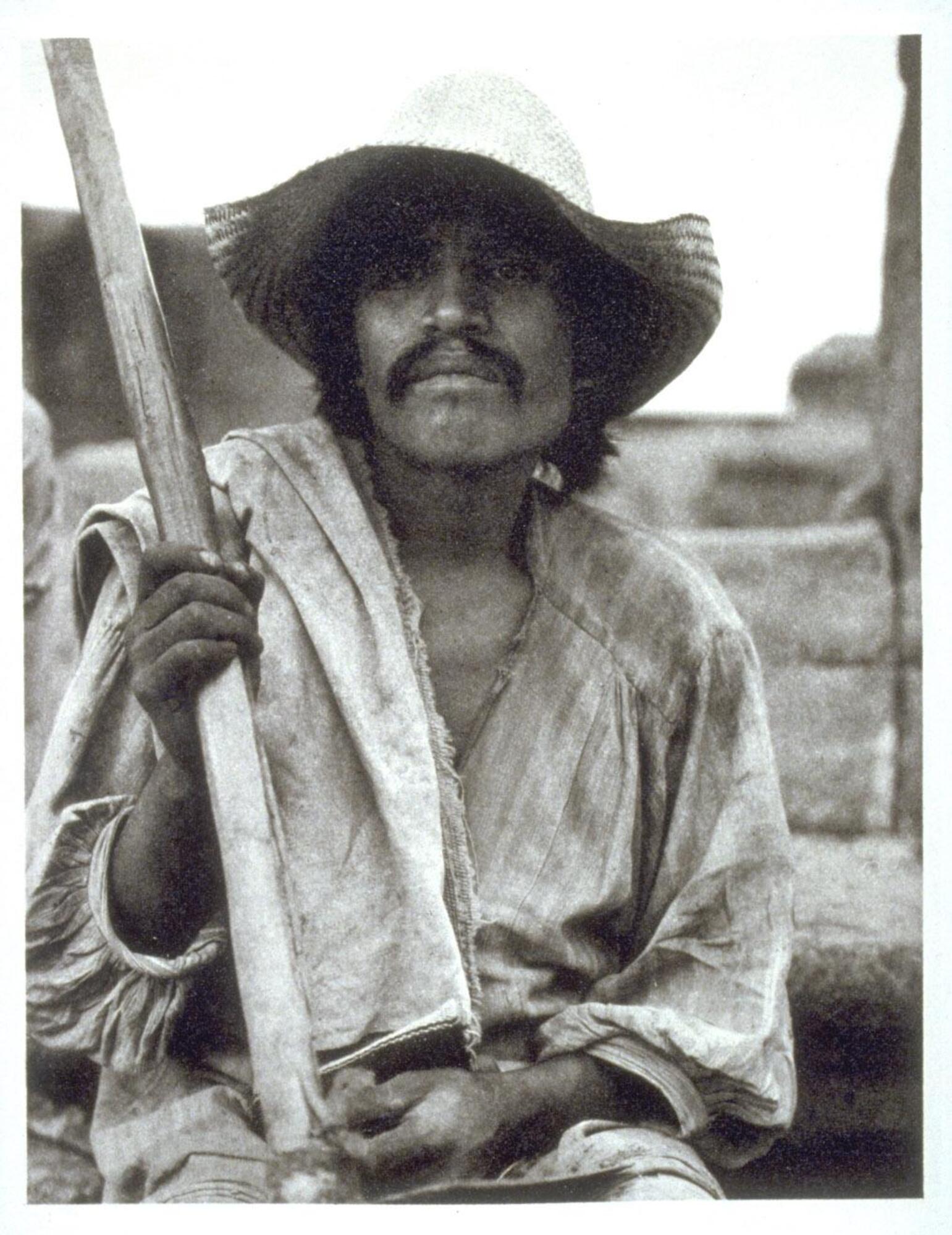 Portrait of a man holding a hoe, wearing soiled campesino clothing. He sits in front of a stone wall, looking out of frame. 