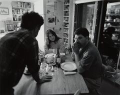 A photograph of a man and woman seated at a dining table, empty plates in front of them. A third man in the foreground leans toward them in conversation.