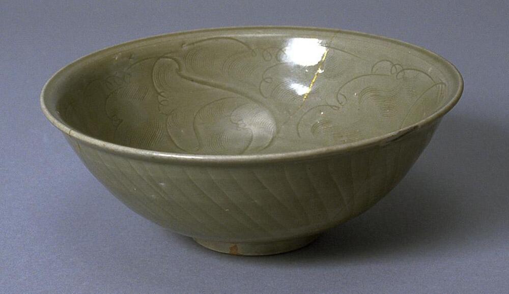 A round stoneware bowl on a footring with slightly everted rim.  It is carved on the outside to resemble lotus flowers and incised on the inside with floral meander.  It is covered in a gray-green celadon glaze, with gold lacquer repair.