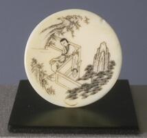A circular ivory snuff dish with a painted ink design of a man in robes sitting on a balcony overlooking trees and mountains.