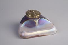 An iridescent glass inkwell with a gold lid.  The base is purplish-gray while the lid has a ram's head engraved on it.