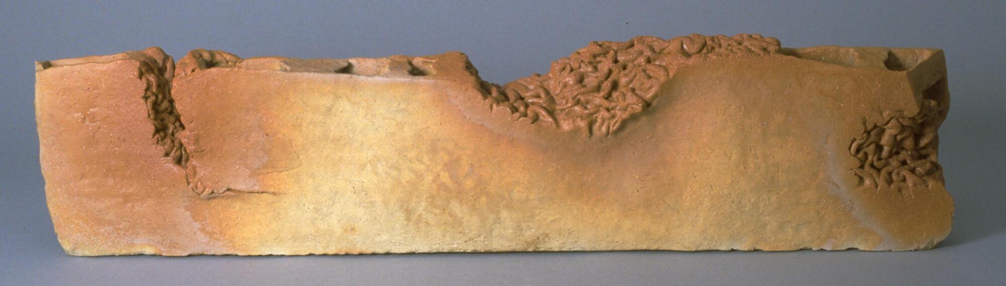 It is a long, rectangular unglazed ceramic piece, intended to be shown in horizontal position. Two thick slabs are connected with bridges inside. The front part has almost flat surface; there is a deep cut on the left side, in which mass of worm-like inner surface can be seen. The same surface is revealed in the middle, as well as on the right edge. The top of the slab has a several shallow holes and one deep cut, inside of which has worm-like surface, as explained above. There is also a dent on the top and on the right, from which worm-like mass seems to be coming out. Reddish shadows cast on left side, in the middle, and the right. There is a patch of clay on left side near the left cut. The bottom is flat.