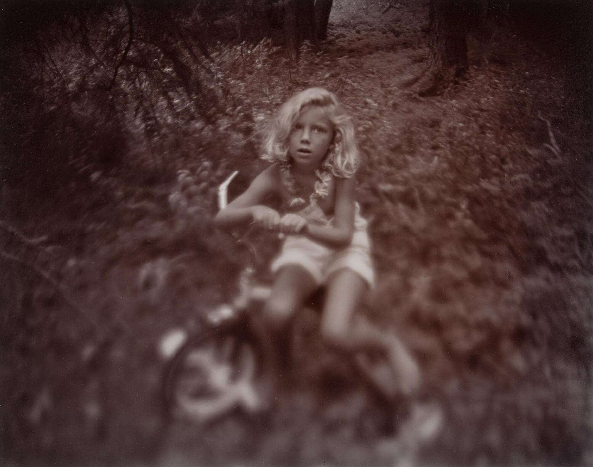 In this photograph of a young girl in a forest clearing she sits on a bicycle, resting her arms on the handlebars. She wears a necklace made of flowers and a pair of light-colored overall shorts.