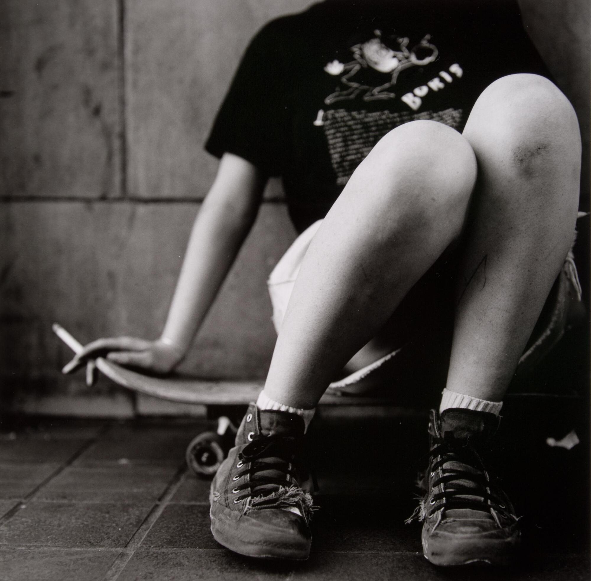 A boy sitting on a skateboard with his head down, smoking a cigarette held in his right hand.